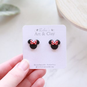 Puede incluir: Un par de pendientes de bot&oacute;n de Minnie Mouse negros con lazos de lunares rojos y blancos. Los pendientes se muestran en una tarjeta blanca con el texto "Ruthie's Art & Clay" y "Handmade Jewelry".