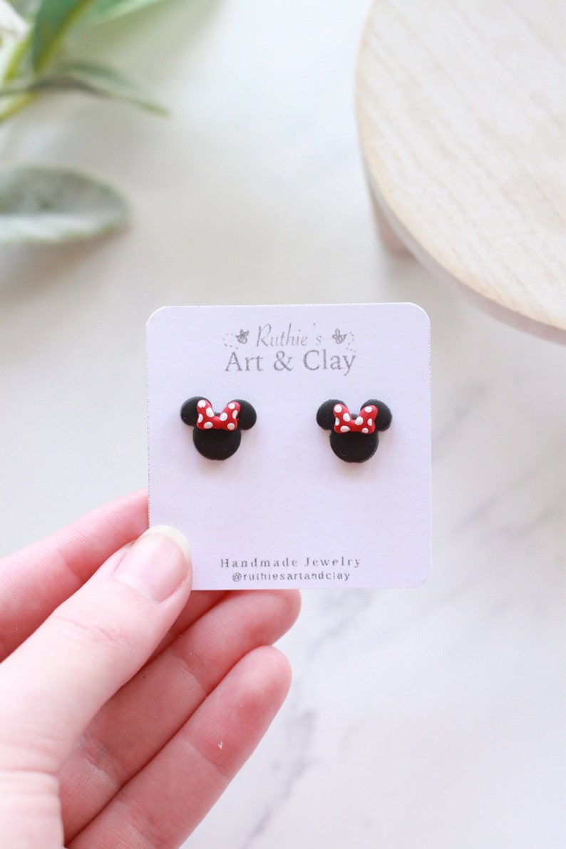 Puede incluir: Un par de pendientes de bot&oacute;n de Minnie Mouse negros con lazos de lunares rojos y blancos. Los pendientes se muestran en una tarjeta blanca con el texto "Ruthie's Art & Clay" y "Handmade Jewelry".