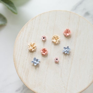 May include: A collection of small, pastel-colored flower embellishments arranged on a light wooden surface. The flowers are in shades of pink, blue, and cream, and are likely used for crafting or decorative purposes. The arrangement is on a round, light-colored wooden stand.
