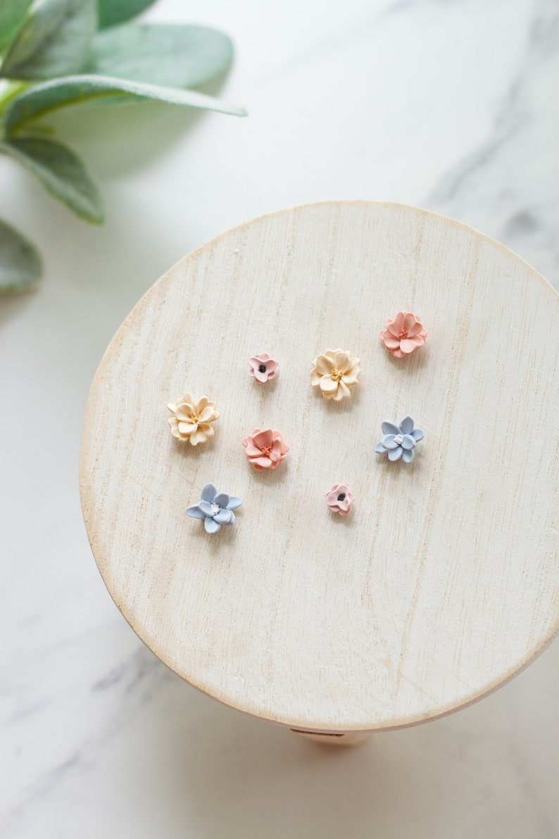May include: A collection of small, pastel-colored flower embellishments arranged on a light wooden surface. The flowers are in shades of pink, blue, and cream, and are likely used for crafting or decorative purposes. The arrangement is on a round, light-colored wooden stand.