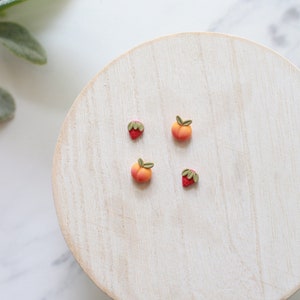 May include: Four miniature fruit-shaped earrings: two peaches and two strawberries. The peaches are orange and yellow with green leaves. The strawberries are red with green leaves. The earrings are displayed on a light wooden surface.
