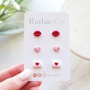 Puede incluir: Una tarjeta con seis pares de pendientes. Los pendientes tienen forma de labios rojos, corazones rosas y sobres blancos con corazones rojos. La tarjeta tiene el texto "Ruthie.Co" y iconos de redes sociales.