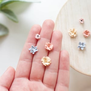 May include: A collection of miniature, handcrafted flower embellishments in various pastel colors, including pink, blue, and yellow. The flowers are arranged on a hand and a wooden surface, showcasing their delicate details and textures.