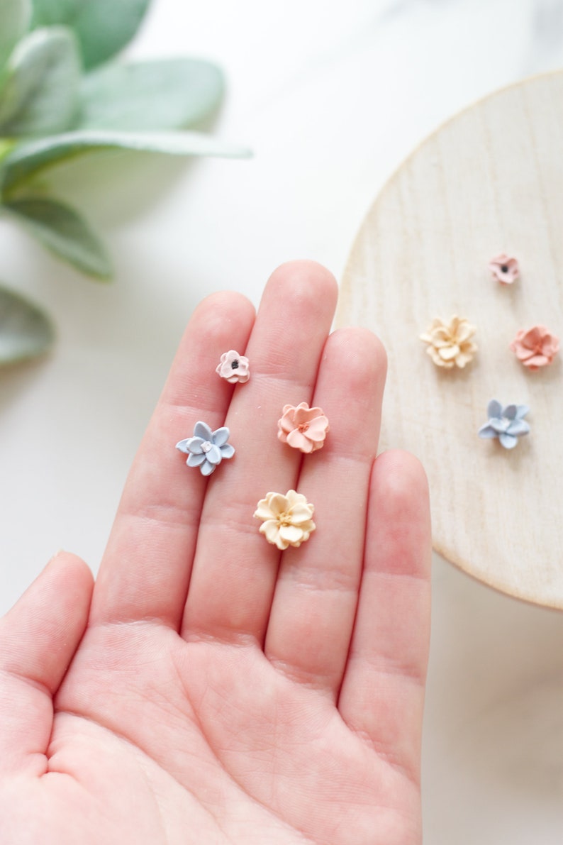 May include: A collection of miniature, handcrafted flower embellishments in various pastel colors, including pink, blue, and yellow. The flowers are arranged on a hand and a wooden surface, showcasing their delicate details and textures.