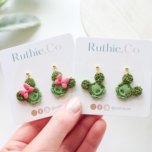 May include: Two pairs of green and pink Minnie Mouse-shaped earrings on white cards. The earrings have a textured appearance and gold-coloured posts. The cards have the text "Ruthie.Co" and social media icons.