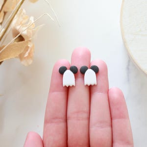 Peut inclure: Une paire de boucles d'oreilles fant&ocirc;mes faites &agrave; la main. Chaque boucle d'oreille pr&eacute;sente un corps de fant&ocirc;me blanc avec un bas festonn&eacute; et une t&ecirc;te circulaire noire. Les boucles d'oreilles sont pr&eacute;sent&eacute;es sur les doigts d'une personne.