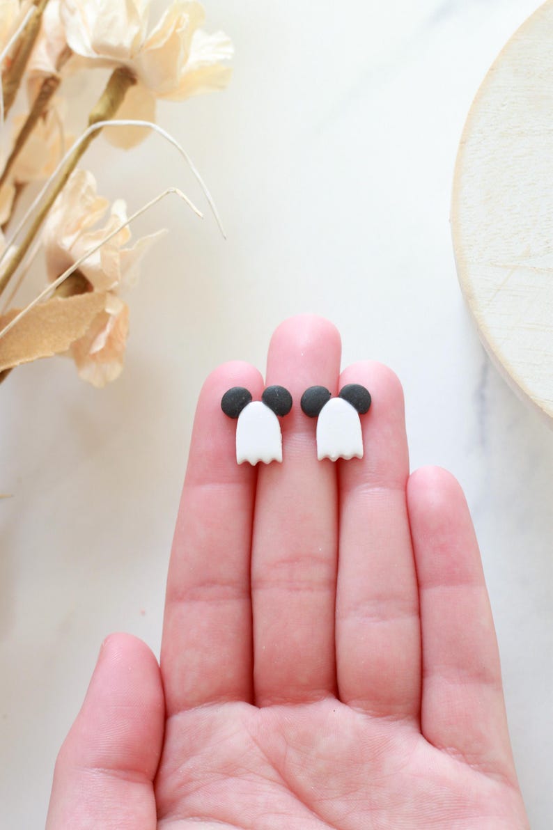 Peut inclure: Une paire de boucles d'oreilles fant&ocirc;mes faites &agrave; la main. Chaque boucle d'oreille pr&eacute;sente un corps de fant&ocirc;me blanc avec un bas festonn&eacute; et une t&ecirc;te circulaire noire. Les boucles d'oreilles sont pr&eacute;sent&eacute;es sur les doigts d'une personne.