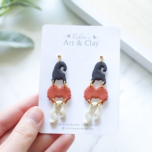 May include: A pair of handmade earrings with a whimsical design. Each earring features a black hat, a rust-colored heart shape, and a cream-colored, curly element. The earrings are attached to gold-colored posts. The card reads "Ruthie's Art & Clay".