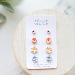 May include: A set of handmade flower stud earrings in various colors, including peach, yellow, and blue, displayed on a white card. The card features the text "Ruthie's Art & Clay" and the Instagram handle @rutiesartandclay.