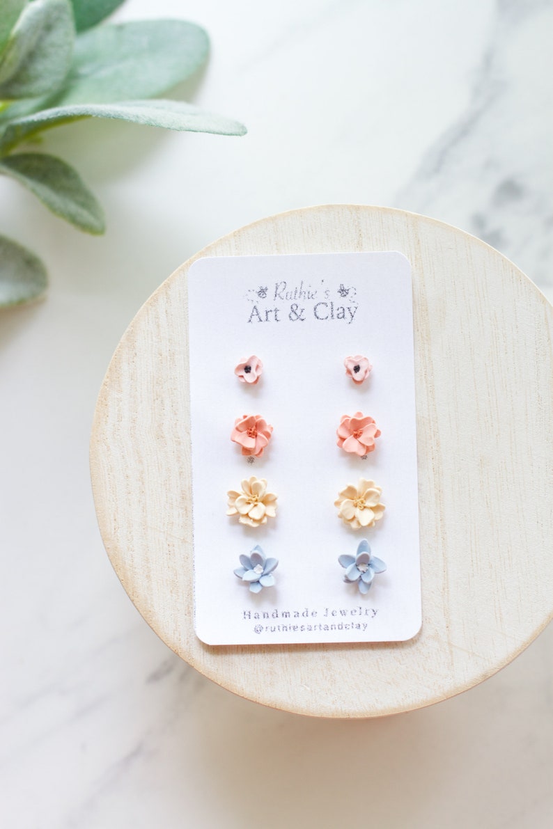 May include: A set of handmade flower stud earrings in various colors, including peach, yellow, and blue, displayed on a white card. The card features the text "Ruthie's Art & Clay" and the Instagram handle @rutiesartandclay.