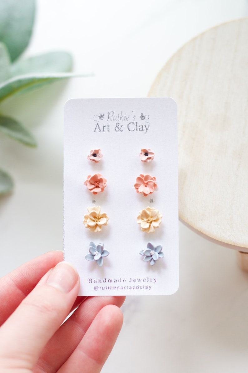 May include: A set of handmade floral stud earrings in various colors, including pink, peach, yellow, and blue. The earrings are displayed on a white card with the text "Ruthie's Art & Clay" and "Handmade Jewelry".