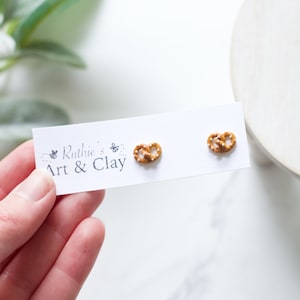Op de afbeelding: Pretzelvormige oorstekers op een witte kaart. De kaart heeft de tekst "Ruthie's Art & Clay". De oorbellen zijn bruin met witte zoutdetails. De achtergrond is wazig wit en groen. De oorbellen zijn waarschijnlijk van klei.