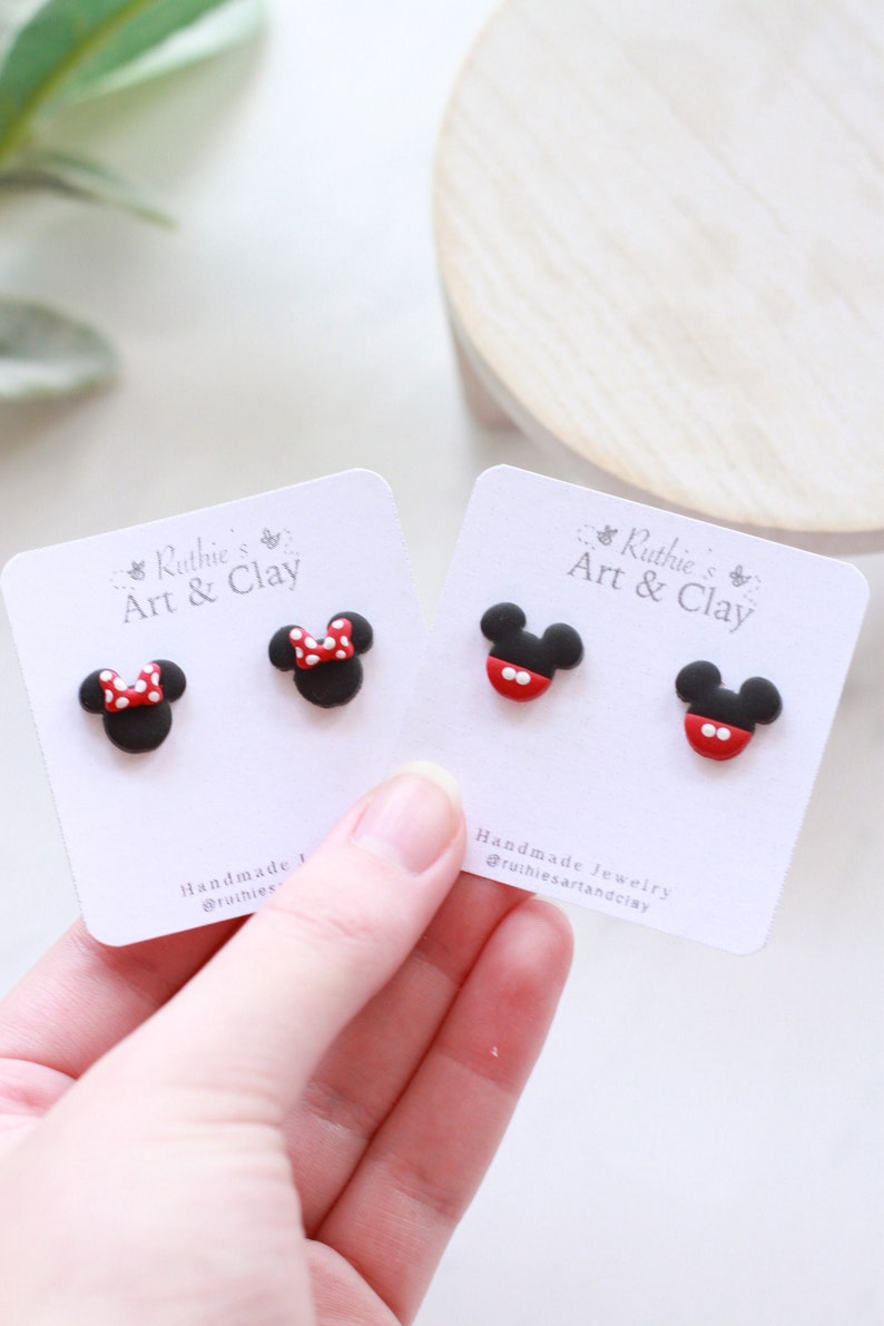 Puede incluir: Pendientes de bot&oacute;n de Mickey Mouse y Minnie Mouse negros y rojos hechos a mano. Un par presenta un lazo rojo con lunares blancos, y el otro un dise&ntilde;o rojo y blanco. Los pendientes est&aacute;n en tarjetas blancas con el texto "Ruthie's Art & Clay".