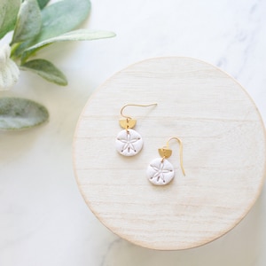 May include: A pair of white sand dollar earrings with gold-colored accents. The earrings are displayed on a light wooden surface, with a marble background and green foliage in the upper left corner.