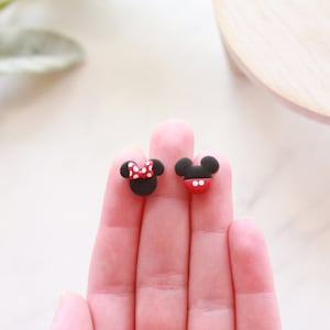 Puede incluir: Dos pendientes negros y rojos con el tema de Mickey Mouse y Minnie Mouse. El pendiente de Minnie Mouse tiene un lazo rojo con lunares blancos. El pendiente de Mickey Mouse tiene una parte inferior roja con dos puntos blancos.