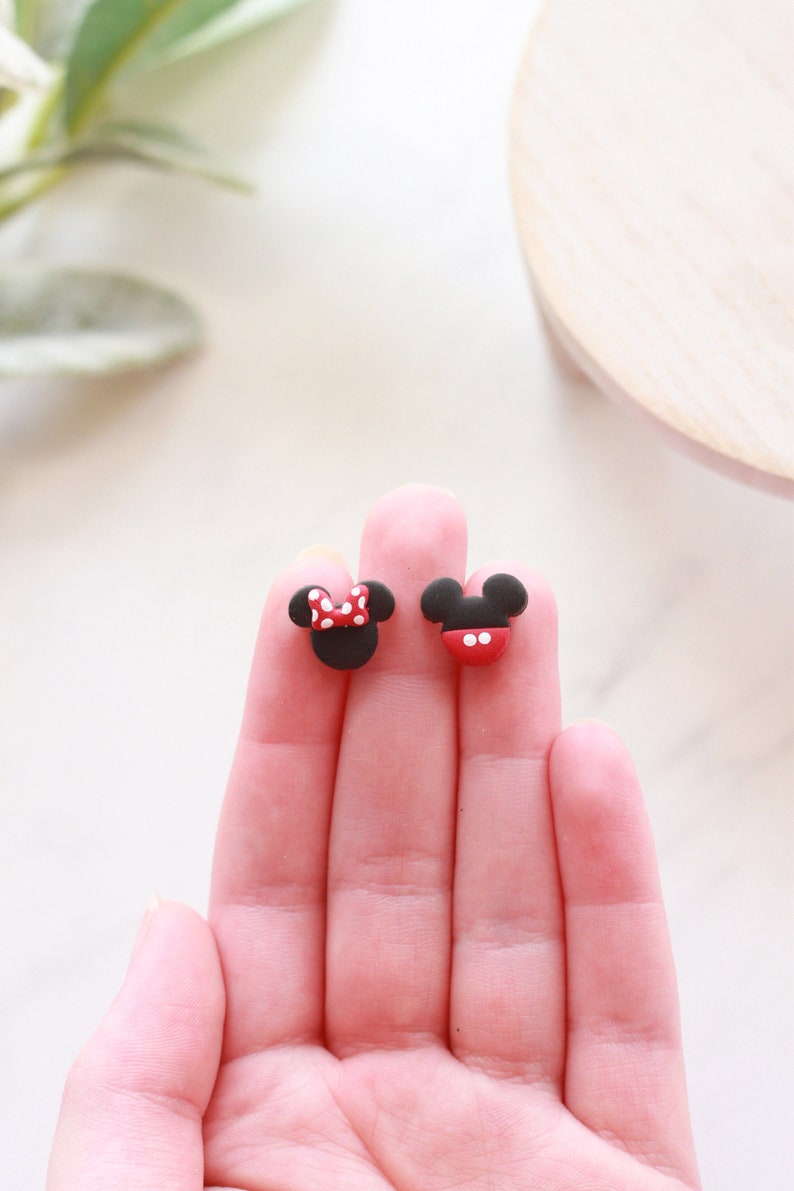 Puede incluir: Dos pendientes negros y rojos con el tema de Mickey Mouse y Minnie Mouse. El pendiente de Minnie Mouse tiene un lazo rojo con lunares blancos. El pendiente de Mickey Mouse tiene una parte inferior roja con dos puntos blancos.