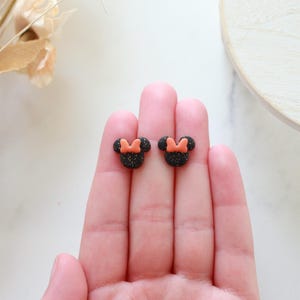Peut inclure: Une paire de boucles d'oreilles &agrave; clous noires paillet&eacute;es en forme de t&ecirc;te de souris avec un n&oelig;ud orange. Les boucles d'oreilles sont petites et sont tenues dans la main d'une personne.