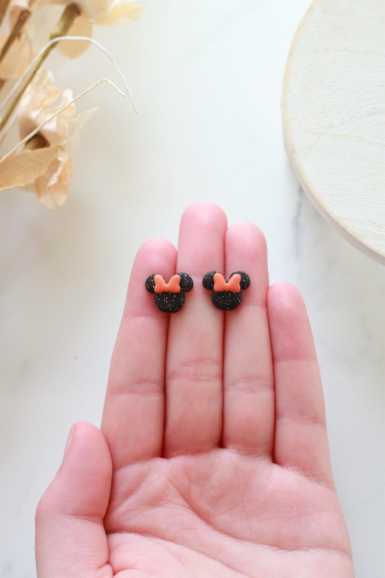 Peut inclure: Une paire de boucles d'oreilles &agrave; clous noires paillet&eacute;es en forme de t&ecirc;te de souris avec un n&oelig;ud orange. Les boucles d'oreilles sont petites et sont tenues dans la main d'une personne.