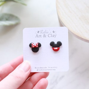 Puede incluir: Pendientes de bot&oacute;n hechos a mano con dise&ntilde;os de Mickey Mouse y Minnie Mouse. Un pendiente tiene un lazo rojo con lunares blancos, el otro una cara roja y blanca. En una tarjeta blanca con el texto "Ruthie's Art & Clay".