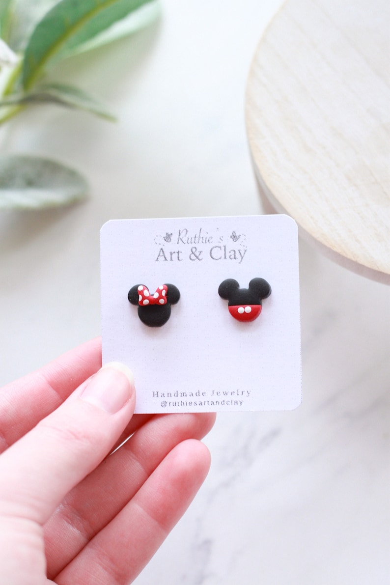 Puede incluir: Pendientes de bot&oacute;n hechos a mano con dise&ntilde;os de Mickey Mouse y Minnie Mouse. Un pendiente tiene un lazo rojo con lunares blancos, el otro una cara roja y blanca. En una tarjeta blanca con el texto "Ruthie's Art & Clay".