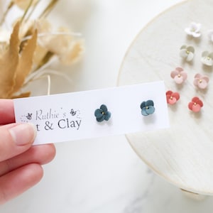Puede incluir: Pendientes de botón de flores hechos a mano en varios colores, incluyendo verde azulado, rosa y blanco, expuestos sobre una superficie blanca. Los pendientes son de arcilla y tienen un diseño floral delicado. La imagen también muestra una tarjeta con el texto "Ruthie's t & Clay."