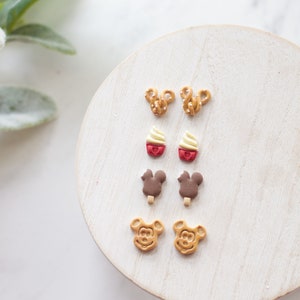 May include: A collection of novelty earrings in various shapes, including pretzels, cupcakes, ice cream bars, and Mickey Mouse faces. The earrings are arranged on a white, round wooden surface, with a sprig of green foliage in the background.