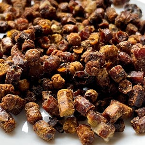 May include: A collection of small, cylindrical bee bread granules in various shades of dark brown, reddish-brown, and golden yellow. These dense, textured pieces have a granular, compressed appearance with irregular shapes and a matte finish.