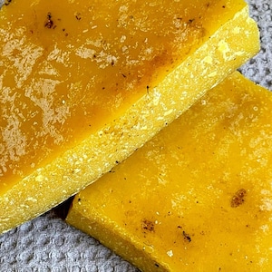 May include: Two blocks of yellow beeswax, one on top of the other. The beeswax has a rough, textured surface.