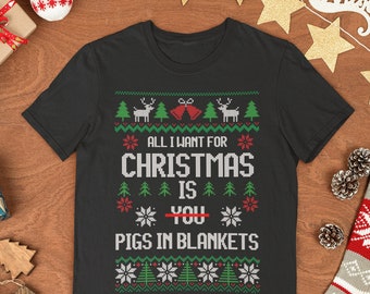 May include: Black t-shirt with a festive Christmas design. The design includes reindeer, Christmas trees, snowflakes, and the text "ALL I WANT FOR CHRISTMAS IS PIGS IN BLANKETS". The word "YOU" is crossed out in red.