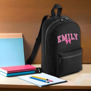 May include: Black canvas backpack with a pink bow and the name "EMILY" embroidered on the front.