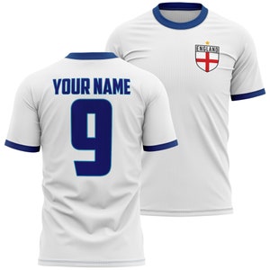 Könnte beinhalten: Weißes Fußballtrikot mit blauen Rändern und dem England-Wappen auf der Vorderseite. Auf der Rückseite des Trikots befindet sich die Nummer 9 und der Text "YOUR NAME" in blau.