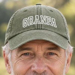 May include: An olive green baseball cap with a curved brim and a structured crown. The word GRANPA is embroidered in white capital letters with a bold outline across the front centre of the cap. The fabric has a slightly weathered, textured appearance.