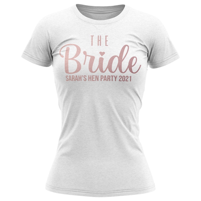 K&ouml;nnte beinhalten: Wei&szlig;es T-Shirt mit dem Text "The Bride" in ros&eacute;goldenen Lettern mit einem kleinen Herzen &uuml;ber dem "e". Der Text "Sarah's Hen Party 2021" befindet sich unter dem Haupttext.