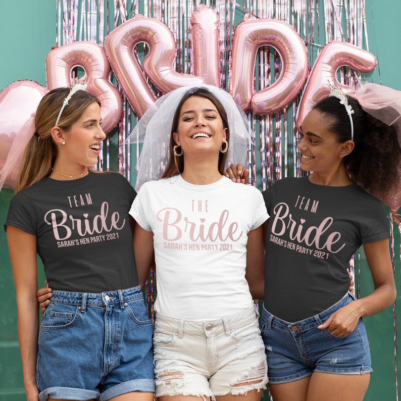 K&ouml;nnte beinhalten: Drei Frauen tragen schwarze und wei&szlig;e T-Shirts mit dem Text "Team Bride" und "The Bride" in rosa Schrift. Die Shirts enthalten auch den Text "Sarah's Hen Party 2021".