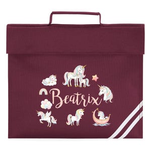 May include: Burgundy school satchel featuring a handle and a flap closure. The front displays a collection of unicorn illustrations with pastel manes and a star. The name "Beatrix" is written in cursive. White stripes run down the right side.