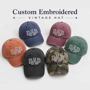 May include: Six baseball caps in various colors including blue, maroon, orange, black, green, and camouflage. Each cap features white embroidered text that reads DAD EST. 2027 in a collegiate font style. The hats have a washed, worn-in appearance.