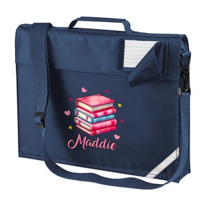 May include: A navy blue school satchel with a shoulder strap and a top handle. It has a graphic of stacked books in pink and blue, with hearts and stars. The name "Maddie" is written in pink script.