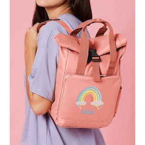 May include: A pink backpack with a rainbow and clouds embroidered on the front. The name "Amira" is embroidered below the rainbow.