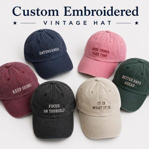May include: Six baseball caps in navy, pink, maroon, black, beige, and dark green, each featuring embroidered text: DAYDREAMER, GOOD THINGS TAKE TIME, KEEP GOING, FOCUS ON YOURSELF, IT IS WHAT IT IS, and BETTER DAYS AHEAD. The hats have a vintage finish.