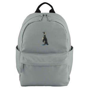 May include: A grey backpack with a black handle and two side pockets. The front of the backpack features an embroidered penguin design in black, white, and yellow. The penguin is standing on a blue base.
