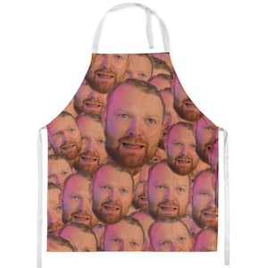 Könnte beinhalten: Eine weiße Schürze mit einem sich wiederholenden Muster eines Männergesichts. Der Mann hat einen Bart und lächelt.