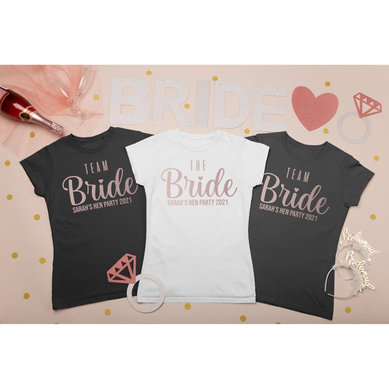 K&ouml;nnte beinhalten: Drei T-Shirts mit dem Text "Bride" in rosa Schrift. Die Shirts sind schwarz, wei&szlig; und grau. Der Text auf den Shirts lautet "Team Bride Sarah's Hen Party 2021" und "The Bride Sarah's Hen Party 2021".