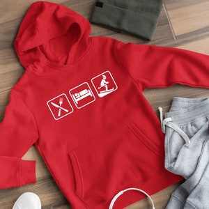 Op de afbeelding: Rood hoodie met een witte afbeelding van drie pictogrammen: een vork en mes, een bed en een skiër. De tekst "Eten, Slapen, Skiën" is impliciet.