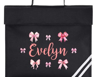 May include: Black school satchel with a top handle and a flap closure. The satchel is decorated with pink bows and the name "Evelyn" in a cursive font. White stripes run down the right side of the bag.