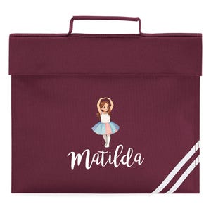 May include: A burgundy school satchel with a handle and a flap closure. The front features a graphic of a ballerina and the name "Matilda" in white script. White stripes accent the lower right corner.