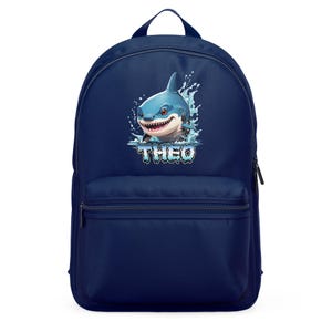 May include: A navy blue backpack with a cartoon shark illustration. The shark is blue and white, with a smiling face and the name "THEO" in blue. The backpack has a front pocket and a top handle.
