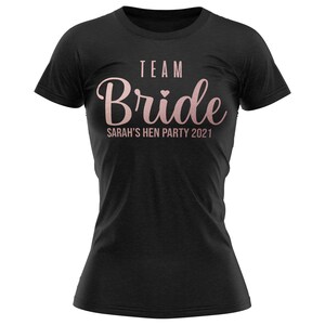 K&ouml;nnte beinhalten: Schwarzes T-Shirt mit dem Text "TEAM Bride" in ros&eacute;goldenen Lettern mit einem kleinen Herz. Der Text "SARAH'S HEN PARTY 2021" befindet sich unter dem Haupttext.