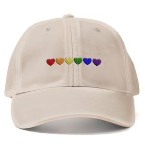 May include: A beige baseball cap with a curved peak. The cap has six embroidered hearts in rainbow colours: red, orange, yellow, green, blue, and purple. The cap has a fabric strap closure at the back.