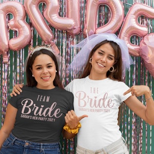 K&ouml;nnte beinhalten: Zwei Frauen tragen passende T-Shirts f&uuml;r eine Junggesellenparty. Die zuk&uuml;nftige Braut tr&auml;gt ein wei&szlig;es T-Shirt mit dem Text "The Bride Sarah's Hen Party 2021" in rosa Schrift. Die andere Frau tr&auml;gt ein schwarzes T-Shirt mit dem Text "Team Bride Sarah's Hen Party 2021" in rosa Schrift.