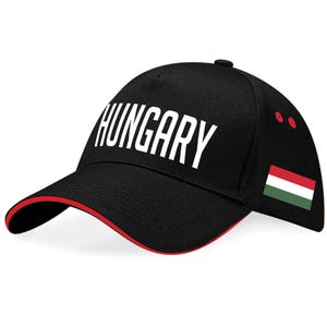 May include: Black baseball cap featuring the word "HUNGARY" in white. The cap has a red trim and the Hungarian flag on the side. Two red dots are on the side of the cap.